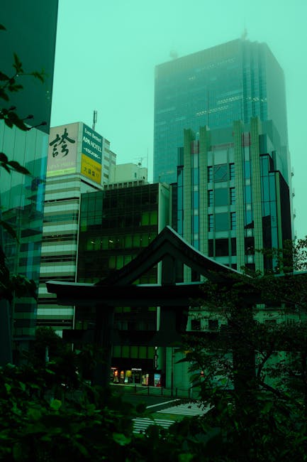 A tall modern glass office building towering above smaller commercial structures in an urban setting, with a traditional Japanese torii gate in the foreground partially obscured by greenery. The scene appears to be taken during daytime with subdued lighting due to overcast weather or mist. The buildings have illuminated signs, including one with Japanese characters and an advertisement for a hotel. The image reflects a blend of contemporary architecture and cultural elements, with no visible furniture or moving activities. This setting highlights the challenges of navigating narrow urban streets during home relocation or furniture transport, relevant to professional removals services like Man with Van Chelsfield, who specialise in logistical planning for such environments.