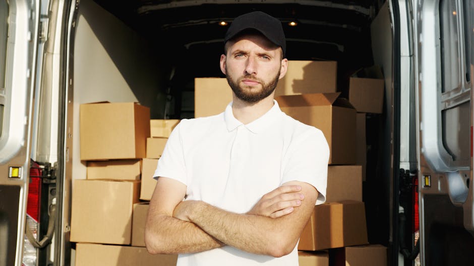 A young man with a beard, wearing a red cap backwards, a checked shirt, and a beige and brown jacket, is sitting on the edge of an open van's loading area during a house removal process. The van's int