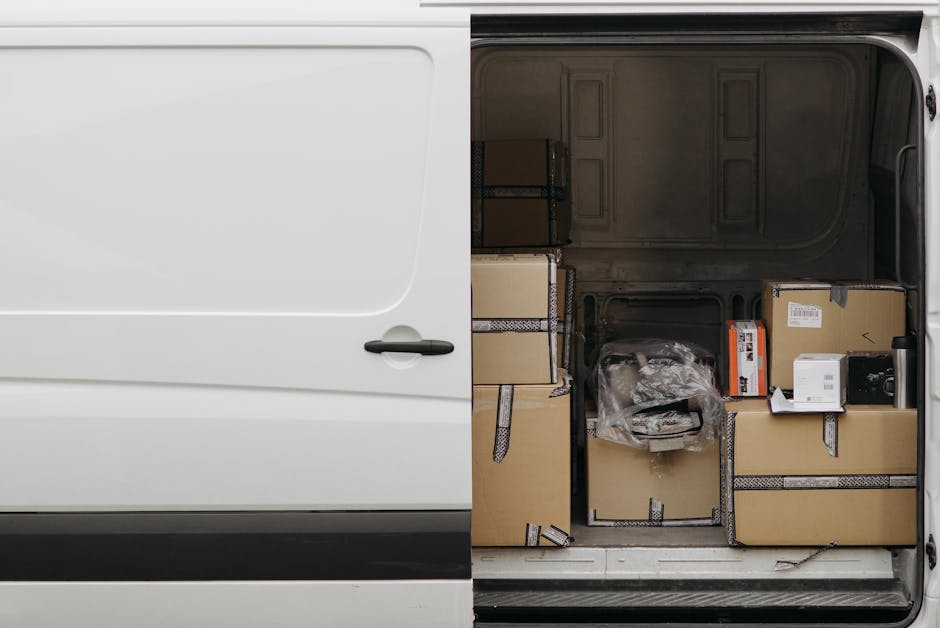 Inside the rear compartment of a van used for home relocation, several cardboard boxes of various sizes are arranged for furniture transport and packing and moving activities. Some boxes are sealed with packing tape, while others are open with plastic wrapping or loose contents visible. The boxes are positioned on the vehicle's floor, with larger boxes placed against the walls and smaller ones stacked nearby. The van's interior features a grey metal lining, and the open rear door reveals an outdoor pavement area, indicating the loading process is underway for a house removal. The environment is well-lit, highlighting the organized arrangement of packing materials and boxes, consistent with professional removals services offered by Man with Van Chelsfield, based on the Bromley Council permits for Chelsfield van loading.