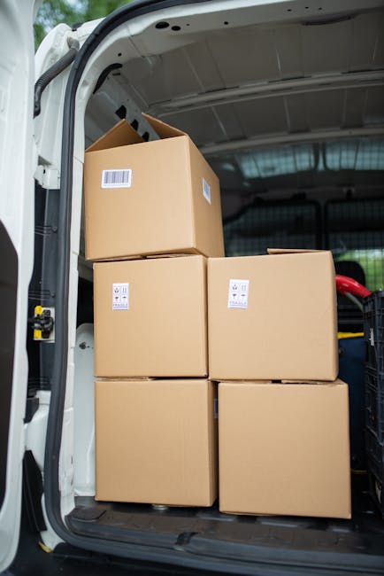 Inside the rear of a white van used for home relocation, five cardboard boxes of varying sizes are stacked neatly, with the topmost box slightly tilted. The boxes are sealed with white packing tape and have barcode labels attached. The van’s interior is well-lit, revealing a dark rubber floor and a black plastic storage compartment on the right side. The surrounding environment outside the van shows a paved loading area, indicating a moving or furniture transport process, likely part of a residential move coordinated by Man with Van Chelsfield, as referenced on the Bromley Council Permits page for Chelsfield van loading.