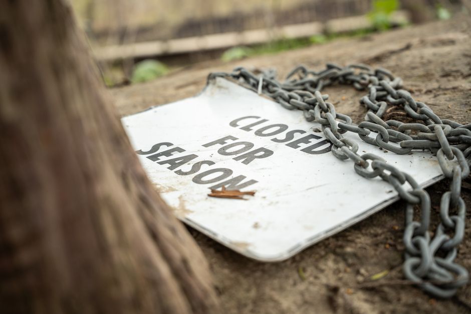 A weathered metal 'Closed for Shushow' sign lying on the ground outside, with a tangled metal chain draped over it. The sign is positioned on a dirt surface near a wooden fence and partially obscured by a tree trunk in the foreground. The background shows a blurred outdoor environment with some green foliage and natural light. The scene suggests an outdoor setting related to house removals or property clearance, with the sign possibly indicating restricted access or waste disposal areas. The presence of the chain and the sign emphasizes safety and regulation compliance, relevant to professional removals or moving logistics handled by company such as Man with Van Chelsfield.