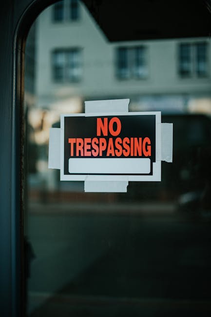 A close-up view of a black and red 'No Trespassing' sign attached to the interior of a vehicle window using white adhesive tape. The sign features bold red text on a black background with a blank white space beneath for additional information. The background shows a blurred exterior scene with buildings and windows, indicating the vehicle is parked outdoors. This image reflects aspects of home relocation or moving processes involving vehicle loading or transfer of belongings, which may be part of a professional removals service such as Man with Van Chelsfield, operating within the context of house removals and transport logistics.