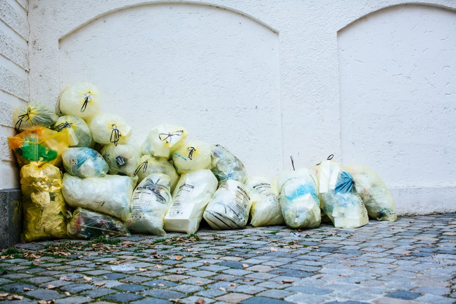A collection of tightly packed rubbish bags, primarily made of yellow and clear plastic, are piled on a cobblestone pavement outside a white exterior wall with a brick corner on the left. The bags contain various household waste items, and some are sealed with black plastic ties. The scene depicts an outdoor waste disposal area, possibly for collection or removal during a home relocation or moving process, as part of a professional service like Man with Van Chelsfield, indicated by the setting and context. The lighting is natural, and the bags are positioned against the wall near a doorway or access point, ready for waste collection or transportation, reflecting typical logistical steps involved in house removals and packing and moving activities.