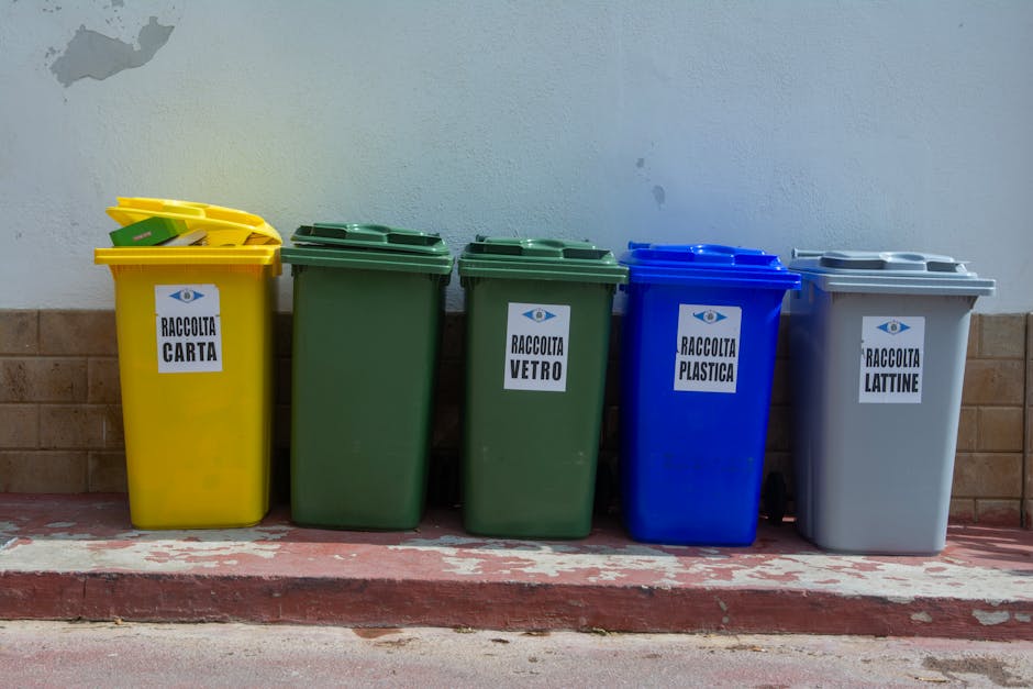 A black and white photograph showing a row of waste disposal bins positioned on a paved area next to a large storage container. The bins include two black bins labeled 'PAPER' and 'CARBOARD ONLY', and four white bins with lids secured by chains. A wooden post is in front of the bins, and a black skip bin or storage container is situated behind them. The background features dense foliage and trees, with a partially visible building roof at the top edge of the image. The scene appears to be part of a residential or commercial property undergoing clearance or waste collection, relevant to house removals and packing logistics, as managed by Man with Van Chelsfield.