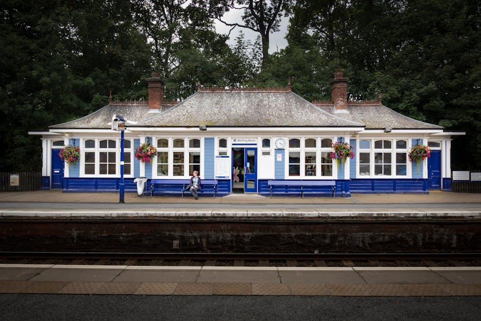 A vintage railway station building situated on a platform, painted in blue and white, with a gabled roof and red brick chimneys. The facade features large windows with multiple panes, and decorative hanging flower baskets with colorful blooms are installed alongside. Two benches are positioned in front of the building, with a person sitting on one and another standing nearby, possibly waiting for a train. The platform is made of concrete, with a tactile paving strip along the edge for safety. In the foreground, train tracks run parallel to the platform, and in the background, tall trees and dense foliage provide a natural backdrop. The image appears to be captured during daytime with diffused natural lighting, giving a clear view of the station's architectural details and the surrounding environment. This scene relates to house removals and moving logistics by illustrating a typical train station setting, where objects such as luggage or boxes may be transported as part of a home relocation process, with [COMPANY_NAME] providing professional removals and transport services.