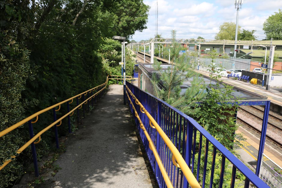 An outdoor pathway adjacent to a railway station with a paved, narrow walkway featuring yellow and blue metal railings on each side. To the left, dense green foliage and trees cast shadows on the pathway, while on the right, the railway lines run parallel, with visible track infrastructure and parked cars in the station car park. In the background, the station platform area with fencing, seating, and overhead electrical wiring is visible under a partly cloudy sky. The scene suggests a typical scene of moving activity, with [COMPANY_NAME] potentially involved in home relocation or furniture transport, though no specific moving actions are visible in this snapshot.
