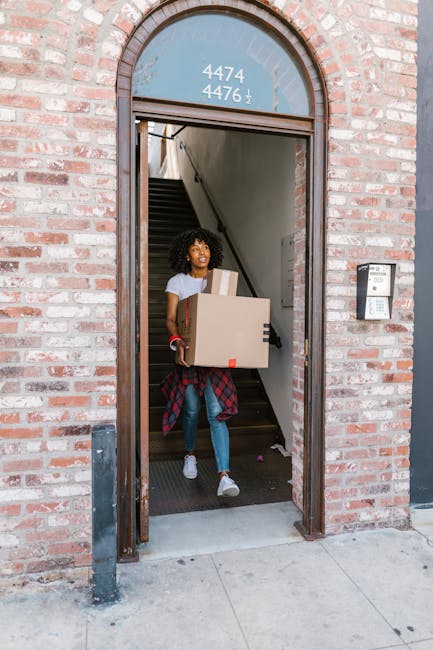 A young woman with curly hair carrying a cardboard box with a smaller box on top, emerging from a brick building's doorway during a home relocation. She is dressed casually in jeans, a t-shirt, and a plaid shirt tied around her waist. The doorway has a rounded top with a blue window displaying white house numbers and is equipped with an intercom system on the right side. Inside, a staircase leading upwards is partially visible, suggesting an interior space ready for moving furniture or boxes. The woman is walking on a concrete pavement outside the building, which is part of a professional removal or moving operation, possibly managed by Man with Van Chelsfield. The scene depicts the packing and transport process typical in house removals services near Chelsfield Station, with supervision of loading and unloading items such as boxes, packaging materials, and possibly small appliances.