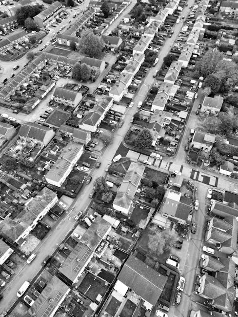 Aerial black-and-white photograph of a residential street featuring terraced houses, semi-detached homes, and small gardens with trees and lawns. Car parks and driveway areas are visible along the street, with numerous parked vehicles, including cars and vans. In the foreground, a section of a street is shown with a few cars parked on the side and a clear paved road. The image captures the dense layout typical of Chelsfield neighbourhood, with closely packed houses, narrow streets, and limited open spaces. This view illustrates the typical environment where professional house removals, such as those provided by Man with Van Chelsfield, may be needed during a home relocation or furniture transport process, with the street serving as a loading area for moving vehicles and equipment.