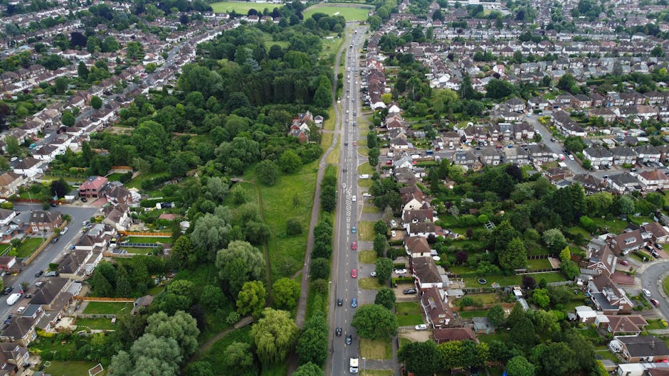 An aerial view of a residential neighbourhood showing tightly packed houses with pitched roofs, some with chimneys, lining a narrow curving street with parked cars along the sides. Adjacent to the street, there is a row of gardens and trees providing greenery. To the right of the houses, a large open grassy area or field extends towards the horizon. The scene is captured during daylight with clear visibility, highlighting the urban layout. In the context of home relocation, this image could relate to the scenic setting of Chelsfield while [COMPANY_NAME], Man with Van Chelsfield, facilitates furniture transport and packing during moving services in the area.