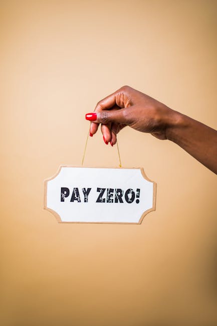 A hand with neatly manicured red nails holds a small white sign with a decorative border, which displays the bold text 'PAY ZERO!' in black capital letters. The hand grips a thin string attached to the top of the sign, suspending it against a plain beige background. The image emphasizes the message of zero cost or free service, relevant to house removals and moving logistics. This visual could relate to promotional offerings from Man with Van Chelsfield, a company specializing in home relocation, furniture transport, and packing and moving services. The sign and hand are captured in close-up, with the background softly blurred, highlighting the sign's message.